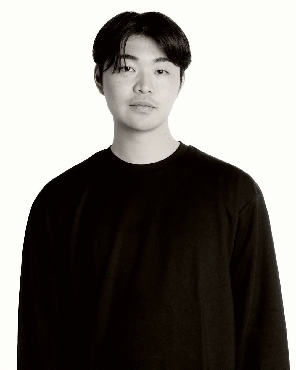 黒の長袖を着て白い壁の前に立つ、KASANE 代表の芦田智。